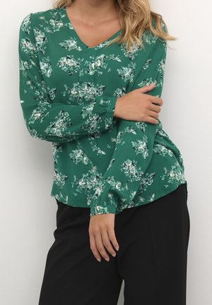 Kobieta ubrana w zieloną, kwiecistą bluzkę z długim rękawem oraz czarne spodnie, pozująca z jedną ręką założoną na drugą na tle gładkiej ściany.