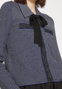 Takkalautanen sinivalkoinen ruudullinen takki mustalla solmiolla, kontrastoiva tekstuuri, koristeelliset napit ja yksityiskohdat taskujen ja alaosan varrella.