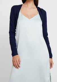 Marinblå cardigan med långa ärmar över en ljusblå, ärmlös klänning med djup V-ringning och mjuk, flödande tyg.
