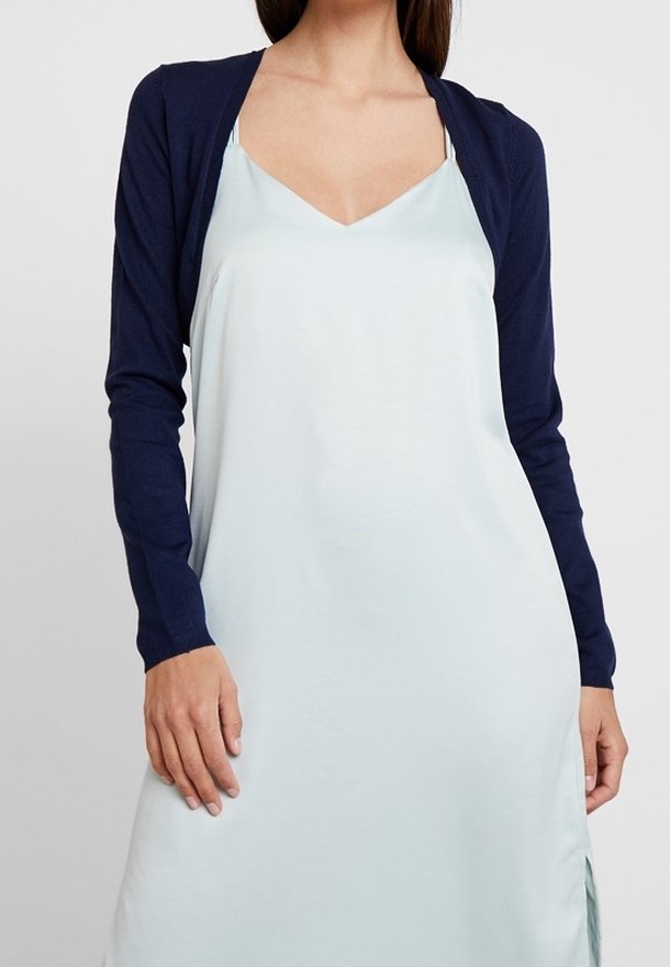 Marinblå cardigan med långa ärmar över en ljusblå, ärmlös klänning med djup V-ringning och mjuk, flödande tyg.