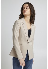 Blazer beige su misura realizzato in tessuto liscio, con chiusura a bottone singolo, revers a punta e tasche laterali. Indossato sopra un top nero.