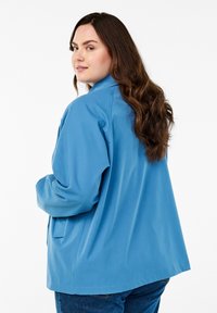 Blå, oversized jacka med en slät textur och spetsig krage, visad bakifrån, ihopparad med mörka jeans.