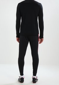 Schwarzes Sportoberteil mit langen Ärmeln und hohem Kragen, kombiniert mit enganliegender schwarzer Hose mit roten Seitenelementen.