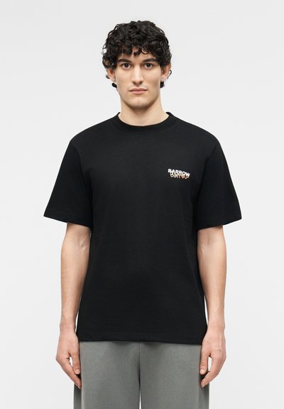 Νεαρός με σγουρά μαλλιά που φοράει μαύρο μπλουζάκι με λογότυπο Barrow και γκρι παντελόνι, στέκεται μπροστά σε απλό ανοιχτόχρωμο φόντο.