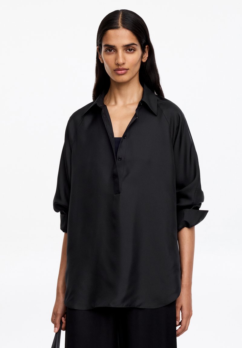 Femme aux longs cheveux foncés portant une chemise noire ample boutonnée avec les manches retroussées, debout devant un fond blanc uni.