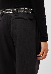 Μαύρο παντελόνι με ζώνη διακοσμημένη με πυραμοειδή καρφιά, πλαϊνές τσέπες και ομαλή υφή, εμφανιζόμενο από το πλάι με χέρι τοποθετημένο στη μέση.