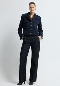 Marineblauwe cropped jas met gouden knopen en twee borstzakken, gecombineerd met hoog-waist donkere spijkerbroeken met zichtbare stiksels en brede pijpen.