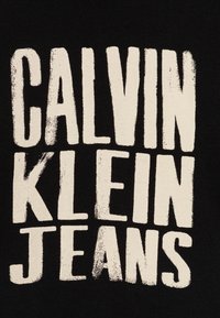Must värvitud kangas, millel on suured, kulunud valged tähet, mis ütlevad "CALVIN KLEIN JEANS." Tähed on julged, ebaühtlaste servade ja erineva paksusega.