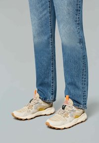 Beige sneakers with orange accents and a textured mesh upper, paired with blue straight-leg jeans. Sole is rubber with a patterned tread.