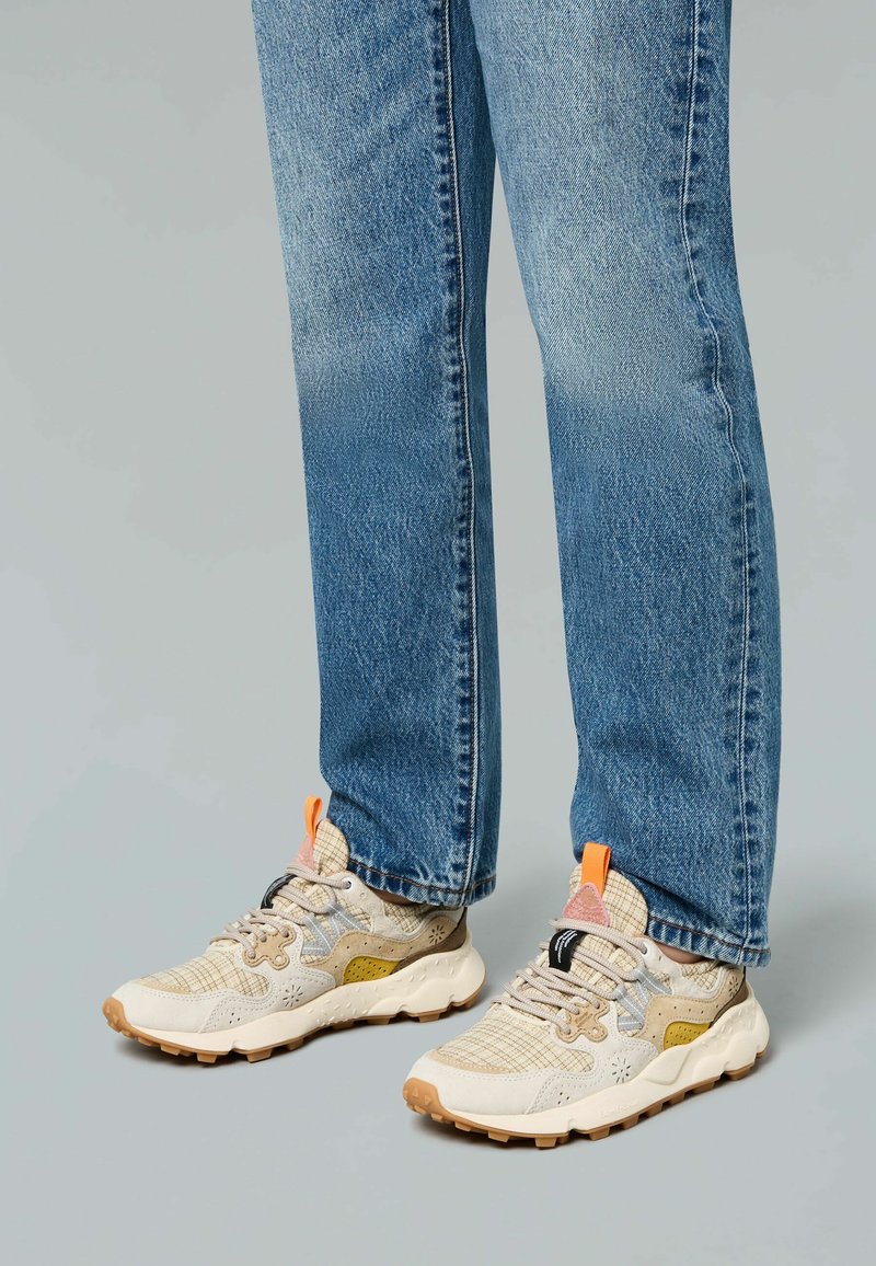 Beige sneakers with orange accents and a textured mesh upper, paired with blue straight-leg jeans. Sole is rubber with a patterned tread.