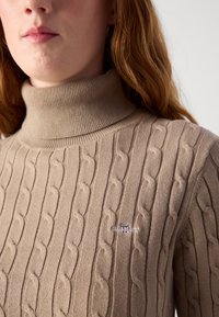 Pull en tricot beige avec col roulé haut, présentant des motifs texturés élaborés et un petit logo sur la poitrine.
