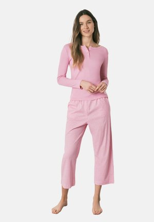 Jeune femme en haut henley rose à manches longues et pantalon large à carreaux vichy rose et blanc, debout pieds nus sur fond blanc.