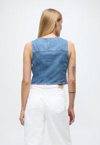 Kratka denim majica s svetlo modrim pranim videzom, brez rokavov, z robom z neobdelanim zaključkom in šivi. Kombinirana z visokim belim hlačami.