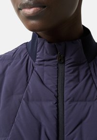 Marineblaue gesteppte Jacke mit hohem Kragen, ausgestattet mit einem Frontreißverschluss und strukturierten Strickakzenten. Der Stoff hat eine glatte, gesteppte Oberfläche.