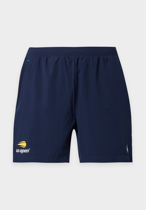 Jeune homme portant un t-shirt Polo blanc, un short bleu marine et des sandales marron foncé, debout devant un fond clair uni.