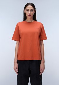 Orange cotton t-shirt with a loose fit and short sleeves, featuring a small embroidered logo in the upper left corner. Black cargo pants.