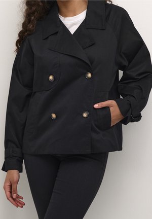 Femme portant une veste noire à double boutonnage avec de gros boutons sur un top blanc, pantalon noir, une main dans la poche, debout devant un fond uni.