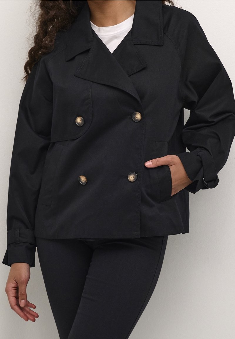 Femme portant une veste noire à double boutonnage avec de gros boutons sur un top blanc, pantalon noir, une main dans la poche, debout devant un fond uni.