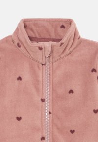 Giacca in pile di un morbido rosa, caratterizzata da un collo alto, una chiusura frontale con zip e motivi di cuori rosa scuro sparsi su tutta la superficie.