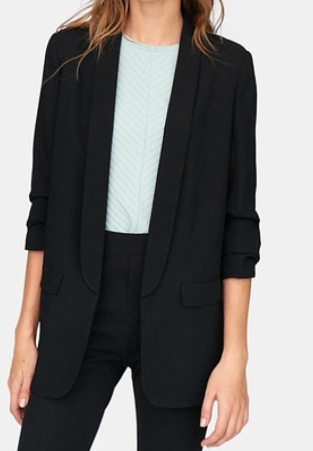 Femme portant un blazer noir aux manches retroussées sur un haut texturé bleu clair et un pantalon noir assorti.