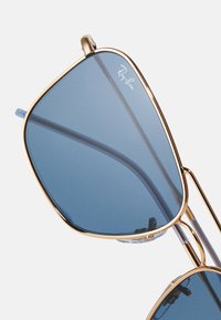 Zlate sončne očala v kovinskem okvirju s plavimi lečami. Imajo edinstveno geometrijsko obliko in nosne blazinice za udobje. Ray-Ban logo na leči.