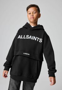 Joven con una sudadera negra con capucha que dice "ALLSAINTS" y "LONDRES", de pie frente a un fondo gris liso.