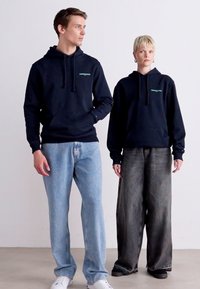 Des sweats à capuche navy avec cordons présentent de petits accents de logo. Le modèle porte un jean baggy bleu clair et gris foncé, complété par des chaussures blanches et sombres.
