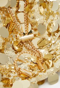 Złota metaliczna naszyjnik z cylindrycznymi wisiorkami, z ogniwami łańcuszka i otoczony rozrzuconymi złotymi cekinami na jasnym tle.