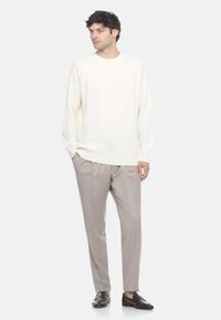 Uomo che indossa un maglione a maglia color crema, pantaloni beige chiaro e mocassini marrone scuro, in piedi con le mani in tasca su uno sfondo bianco.
