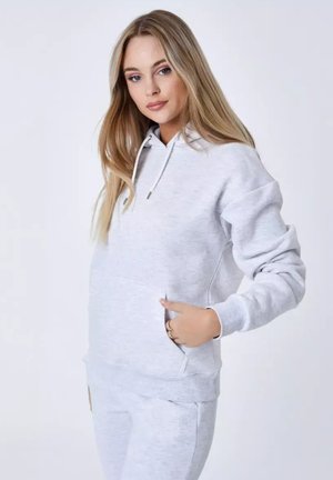 Jeune femme portant un sweat à capuche gris clair et un pantalon de survêtement assorti, debout avec une main dans la poche sur un fond uni.