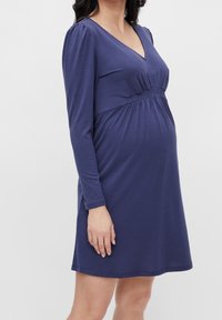 Gravid kvinna som bär en långärmad, marinblå gravidklänning med V-ringning och rynkad midja, som står mot en enfärgad bakgrund.