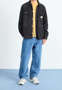 Chemise en denim noir avec deux poches poitrine, associée à un jean baggy bleu clair. Portée avec un T-shirt jaune et des baskets gris-blanc.