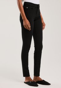 Slim-fit mustad püksid, mida kannab seisev inimene, kelle käed ja jalad on nähtaval, jalas mustad teravate ninadega libisemiskindlad kingad, hele taust.