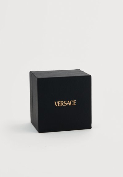 Μαύρο τετράγωνο κουτί με υφή στην επιφάνεια και χρυσό λογότυπο "VERSACE" στο κέντρο της μπροστινής πλευράς, τοποθετημένο σε απλό φωτεινό φόντο.