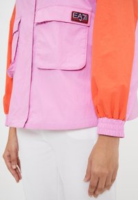 Pink and orange color-block jacket with elastic cuffs, two front pockets, and EA7 logo patch on the chest. Smooth, lightweight fabric.