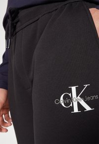 Nahaufnahme von schwarzen Jogginghosen mit dem "Calvin Klein Jeans"-Logo auf dem Oberschenkel und einer Hand, die sich in der Nähe der Tasche befindet.