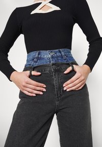 Top negro de manga larga con textura acanalada y un diseño recortado color crema. Combinado con jeans de mezclilla azul desgastado y negro, de talle alto, con las manos en las caderas.