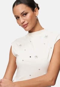 Haut sans manches blanc avec un col montant, orné de motifs de fleurs embellis et de strass dispersés sur l'ensemble du tissu.