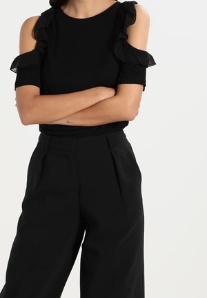 Czarna bluzka z falbanami na odsłoniętych ramionach, zestawiona z czarnymi spodniami o wysokim stanie, ozdobionymi plisowaniem i gładką teksturą tkaniny.