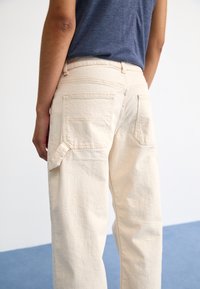 Pantalon crème taille haute en tissu texturé, avec deux poches arrière et un détail de poche latérale, vu de dos.