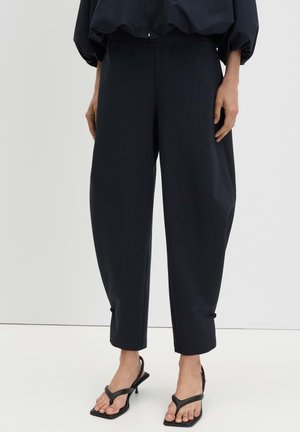Femme portant un pantalon noir ample et des sandales à talons noires, debout sur un sol blanc contre un mur uni.