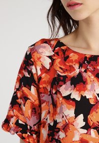 Bloem blouse met een levendig patroon van rood, oranje en roze op een zwarte achtergrond, met een ronde halslijn en korte mouwen.