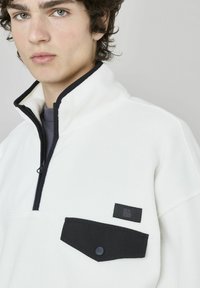 Pull en polaire blanc avec col haut, doté d'une fermeture éclair, de poches noires et d'une patch logo sur la poitrine. Texture douce.