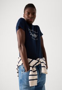 Marineblå bomull T-skjorte med hvit brodert tekst. Lysstripete strikkegenser knyttet rundt livet. Blå denim jeans.