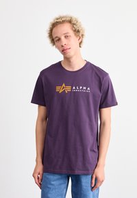 Violet medvilninis marškinėliai trumpomis rankovėmis. Priekyje yra baltas ir oranžinis "ALPHA INDUSTRIES" logotipas ir geometrinių akcentų dizainas.
