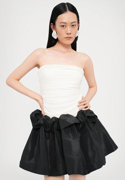 Vestido sin tirantes con parte superior blanca fruncida y una falda negra de satén volumétrica con un dobladillo con volantes. La modelo se encuentra de pie con las manos en las caderas.