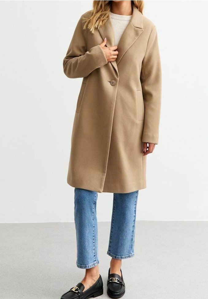 Femme portant un manteau beige à un bouton unique, un pull clair, un jean court bleu et des mocassins noirs avec des boucles dorées, debout devant un fond uni.