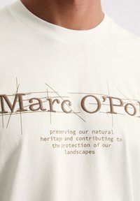 Cremefarbenes Baumwoll-T-Shirt mit gesticktem Text in Braun, das den Markennamen und eine Umweltnachricht zeigt.