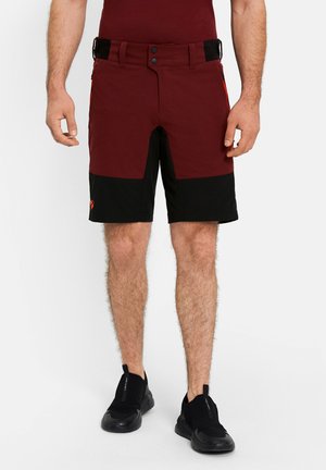 Mann trägt zweifarbige Shorts in Burgunderrot und Schwarz sowie schwarze Slip-On-Sneaker, steht mit locker an den Seiten hängenden Armen.
