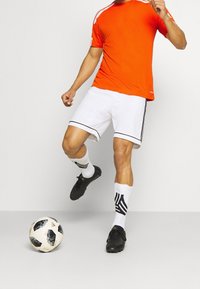 Oranžové sportovní tričko s bílými doplňky, bílé šortky s černým lemováním, černé fotbalové boty a vzorované černobílé ponožky. V záběru fotbalový míč.